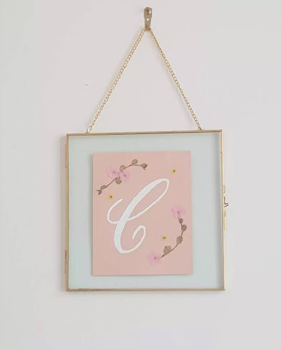 White C letter on a pink background with pressed flowers in a frame